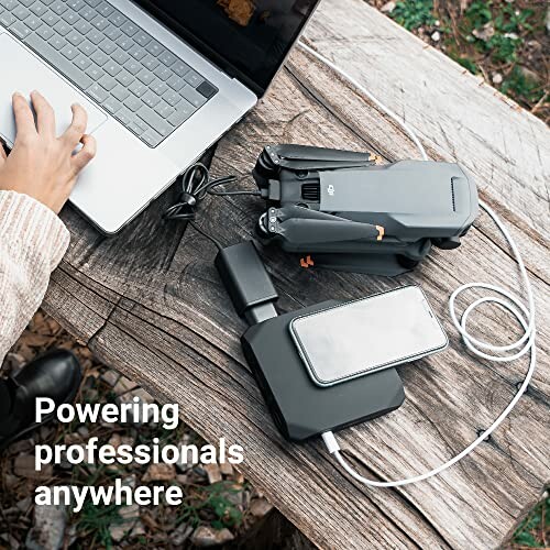 Person using a laptop outdoors with a power bank and drone on a wooden table.